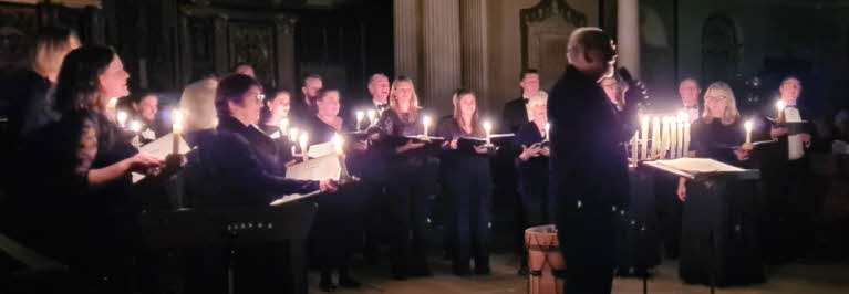 Director Jeffrey Skidmore addressing the audience with the choir standing behind him in a semi-circle, each holding a lit candle.