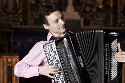 Milos is wearing a pale pink shirt, seated and playing the accordion